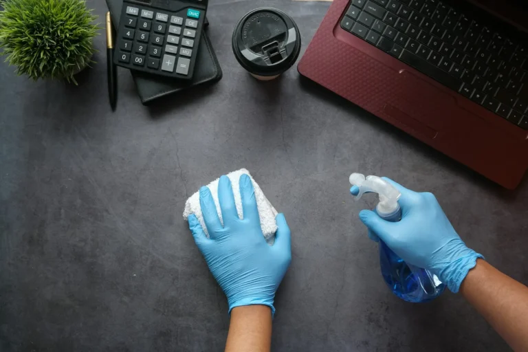 Gloved hands disinfecting an office desk beside a laptop